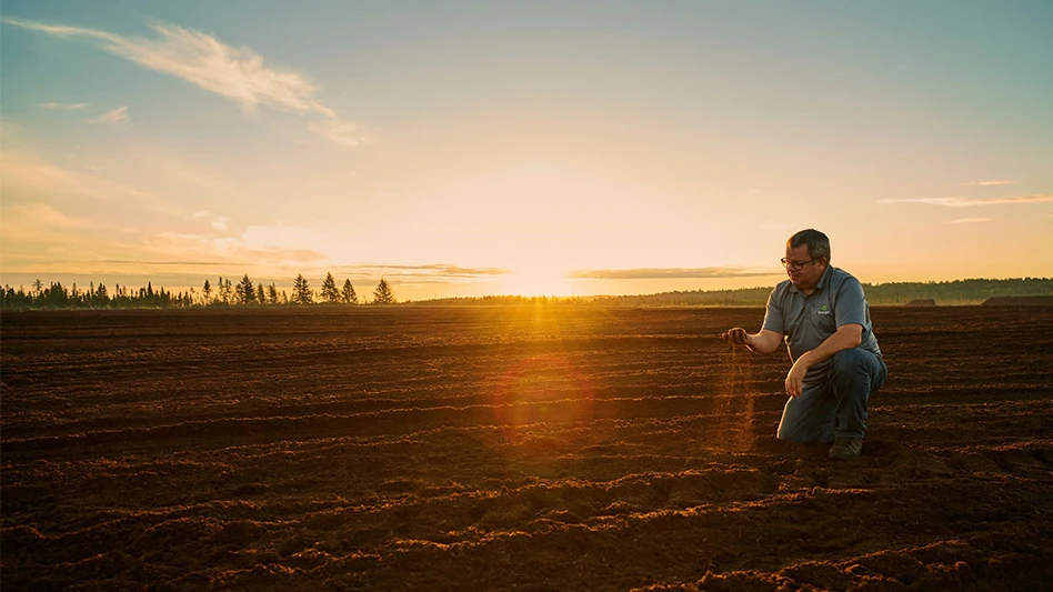 A man wearing a gray polo shirt and dark pants kneels in an empty field with a sunset behind him. He holds a handful of brown soil that falls from his hands.