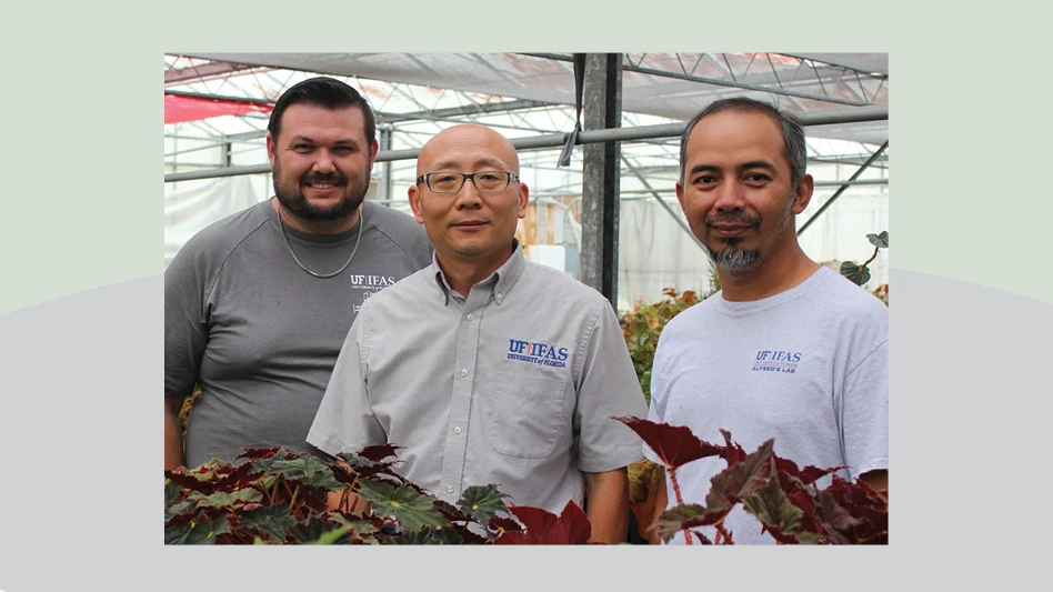 Three men wearing gray shirts pose for a photo in a greenhouse.